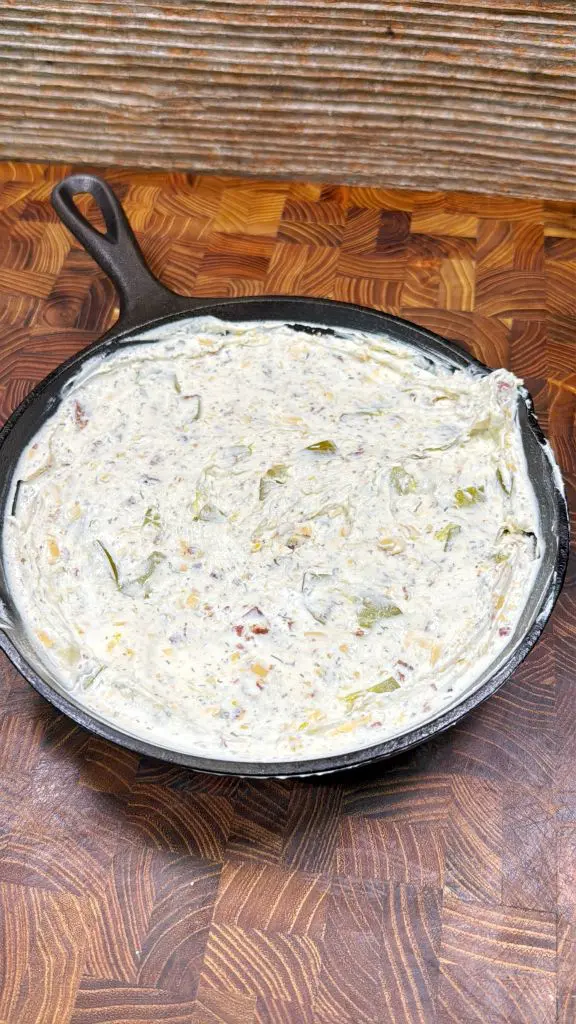 A small cast iron skillet filled with a creamy spinach dip sits on a wooden patterned surface, with swirls and visible green leafy bits in the mixture.
