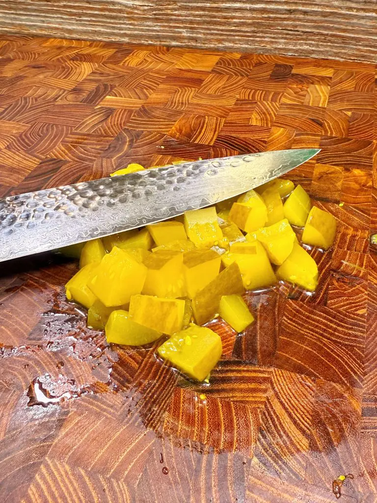 A close-up of a metal knife slicing yellow pickles into small cubes on a patterned wooden cutting board.