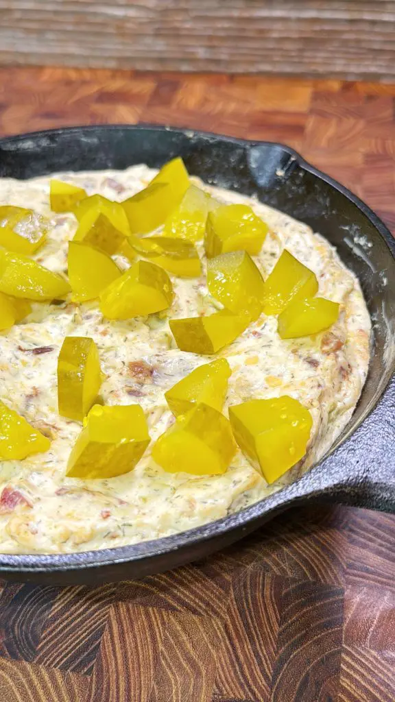 A skillet filled with a creamy dip, topped with chunks of bright yellow pickles, sits on a wooden surface.