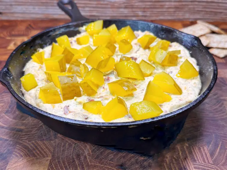 A cast iron skillet filled with creamy dip topped with diced pickles, set on a wooden surface with some crackers nearby.
