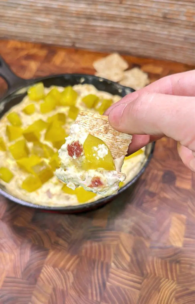 A hand holds a cracker topped with creamy dip, bacon bits, and diced pickles above a skillet filled with more dip and large pickle chunks, on a wooden surface.