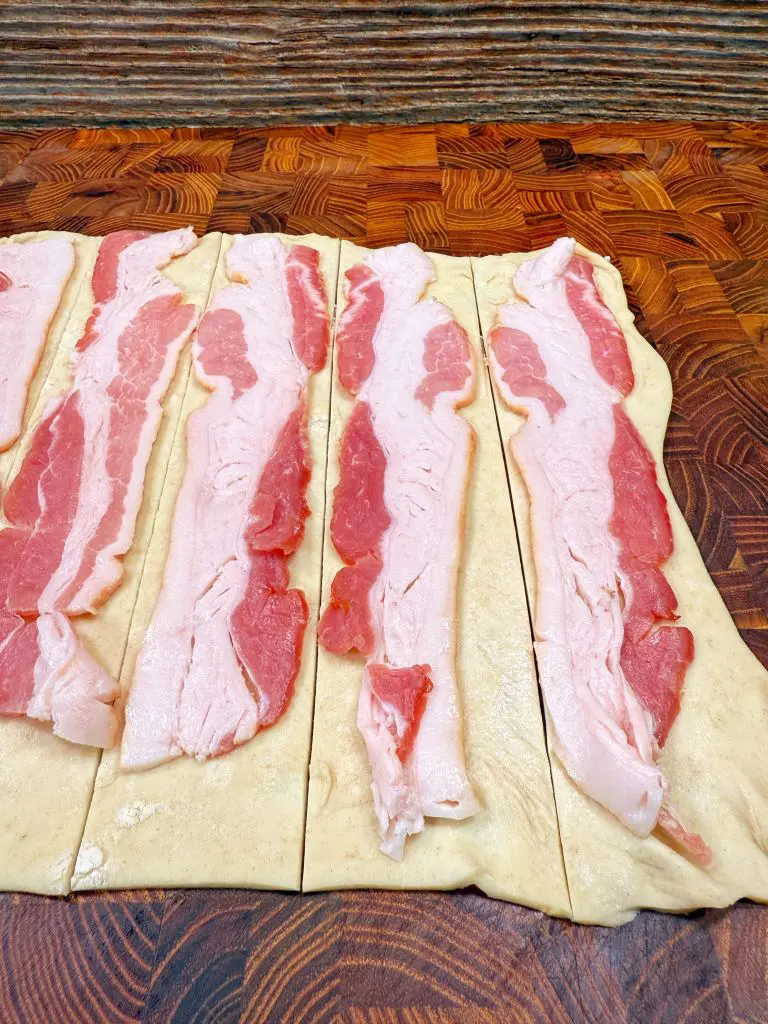 Several raw bacon strips are laid out on rectangular pieces of dough, which are arranged on a wooden cutting board. The surface beneath the cutting board has a striped pattern.