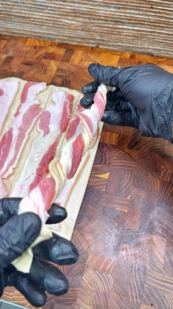 Two hands in black gloves twist a raw strip of bacon over a wooden cutting board with more bacon strips laid out next to it.