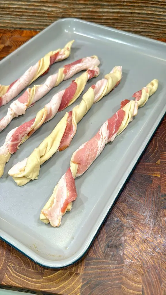 Raw dough strips twisted with uncooked bacon are arranged in rows on a light gray baking sheet, ready to be baked. The baking sheet is on a wooden surface. Recipe is for puff pastry bacon twists.