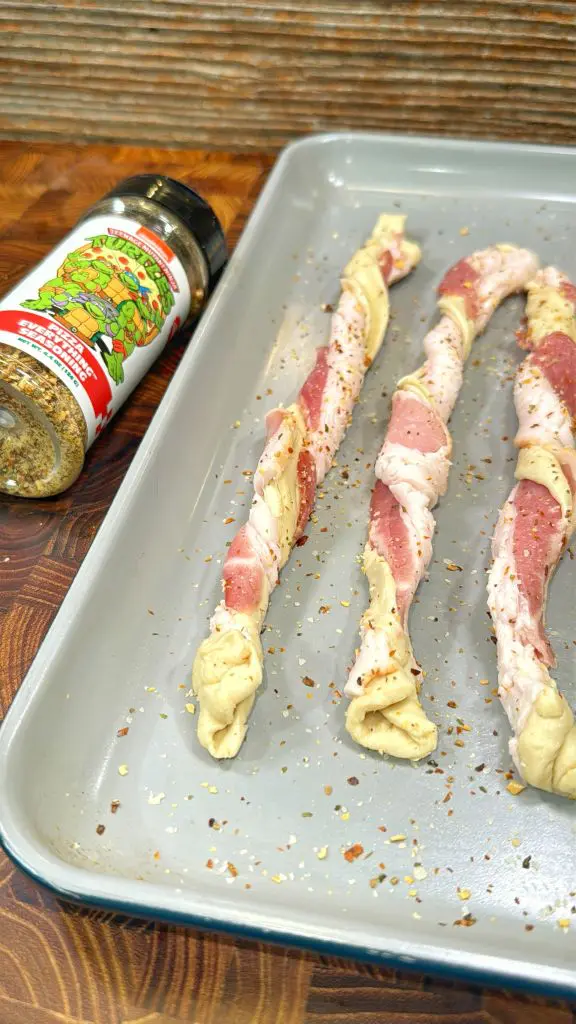 Three raw bacon and dough twists are arranged on a baking sheet, sprinkled with seasoning. An open spice bottle lies nearby on a wooden surface.