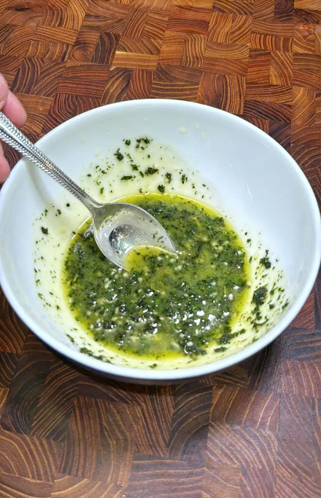A hand holds a spoon in a white bowl containing a green and yellow herb mixture, likely a sauce or marinade, on a wooden surface.