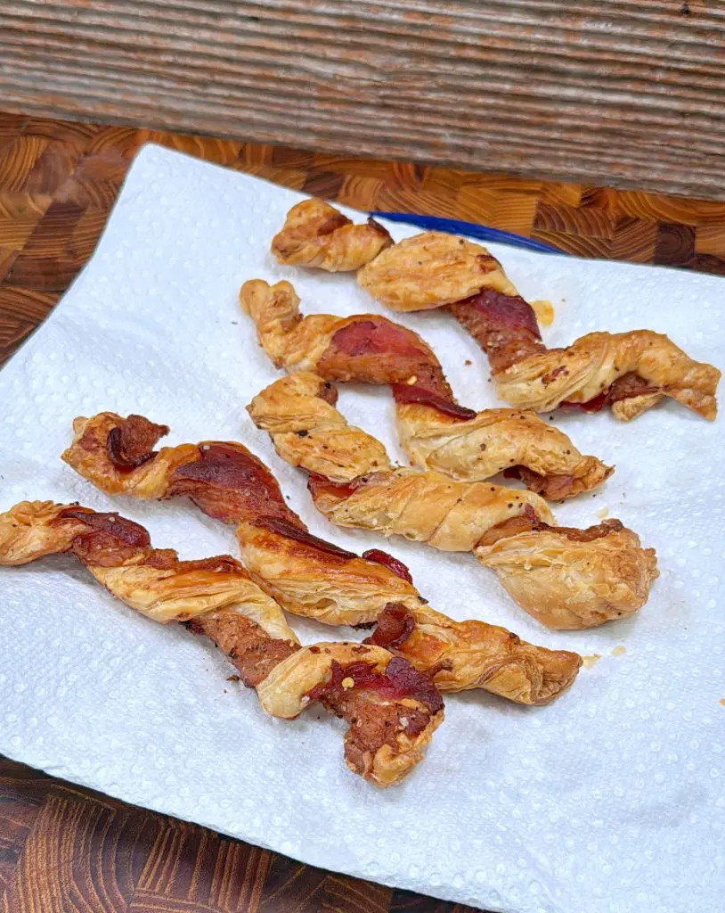 Twisted strips of puff pastry with bacon baked on them are arranged on a white paper towel, placed on a wooden surface with a textured background.