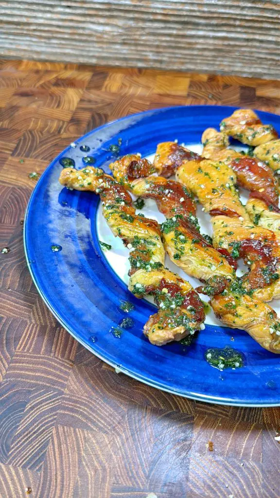 Grilled skewers of meat topped with a green herb sauce are arranged on a vibrant blue plate, set on a wooden patterned surface.
