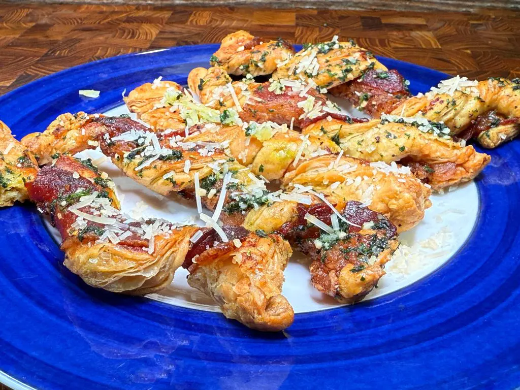 A blue plate holds several puff pastry twists topped with chopped herbs, grated cheese, and visible pieces of pepperoni, displayed on a wood surface.