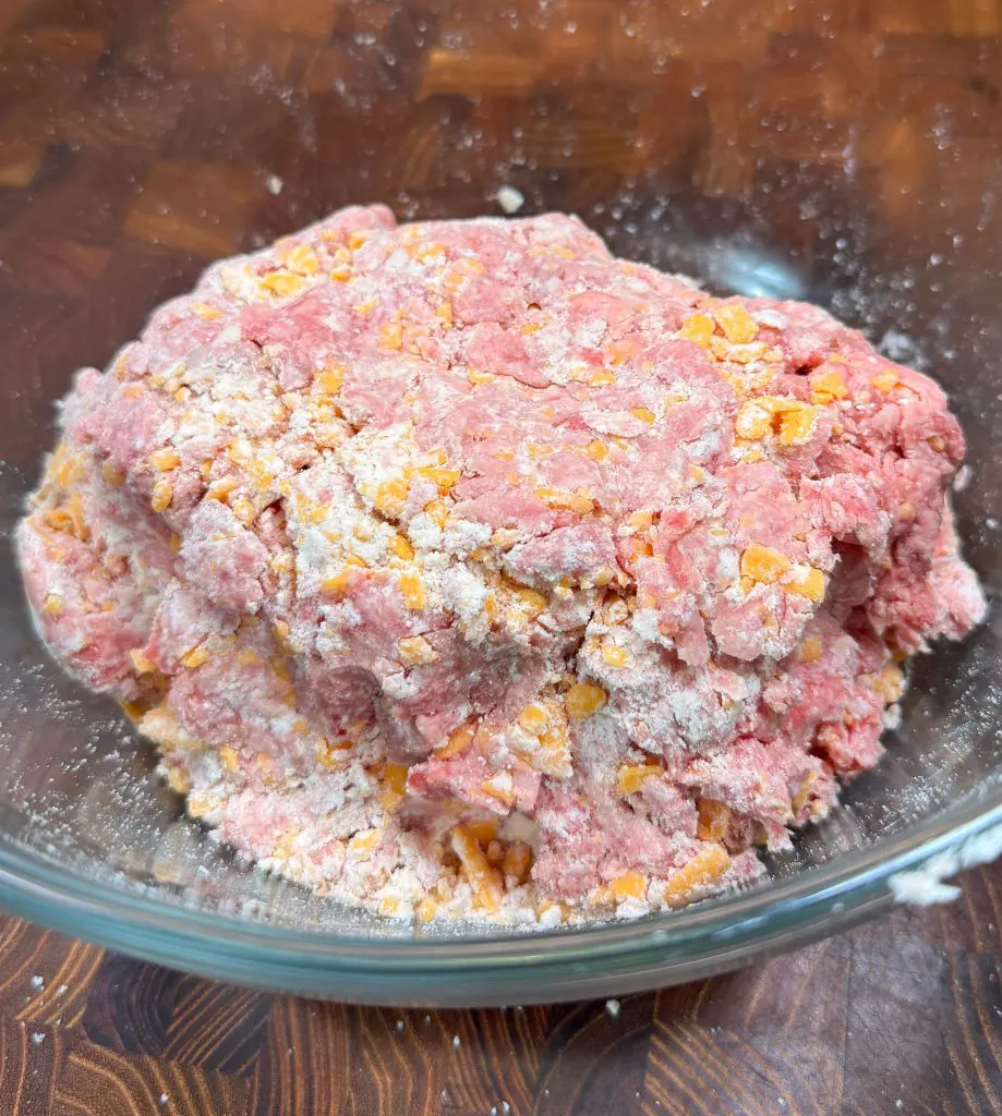 A glass bowl containing a mixture of raw ground meat, shredded orange cheese, and flour on a wooden surface.