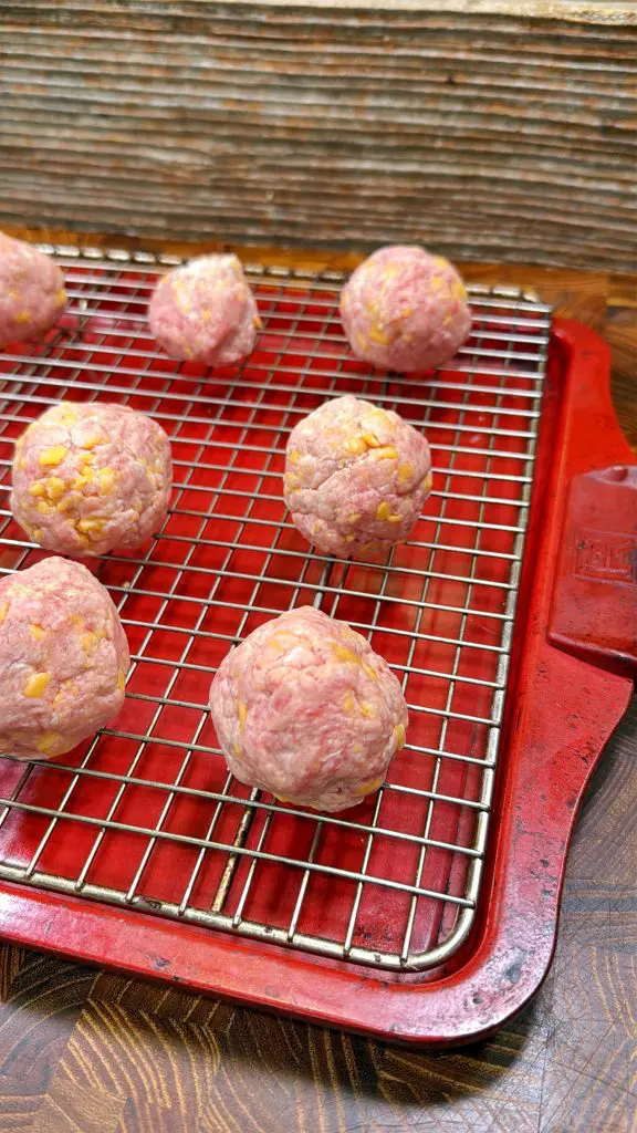 Raw meatballs with visible bits of cheese are arranged on a metal cooling rack placed over a red baking tray, set on a wooden surface.