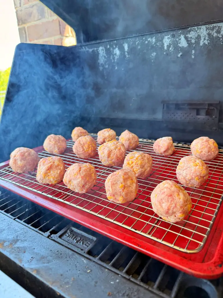 Raw meatballs are arranged on a wire rack over a red tray inside a smoker grill, with smoke rising as they cook. A brick wall and greenery are visible in the background.
