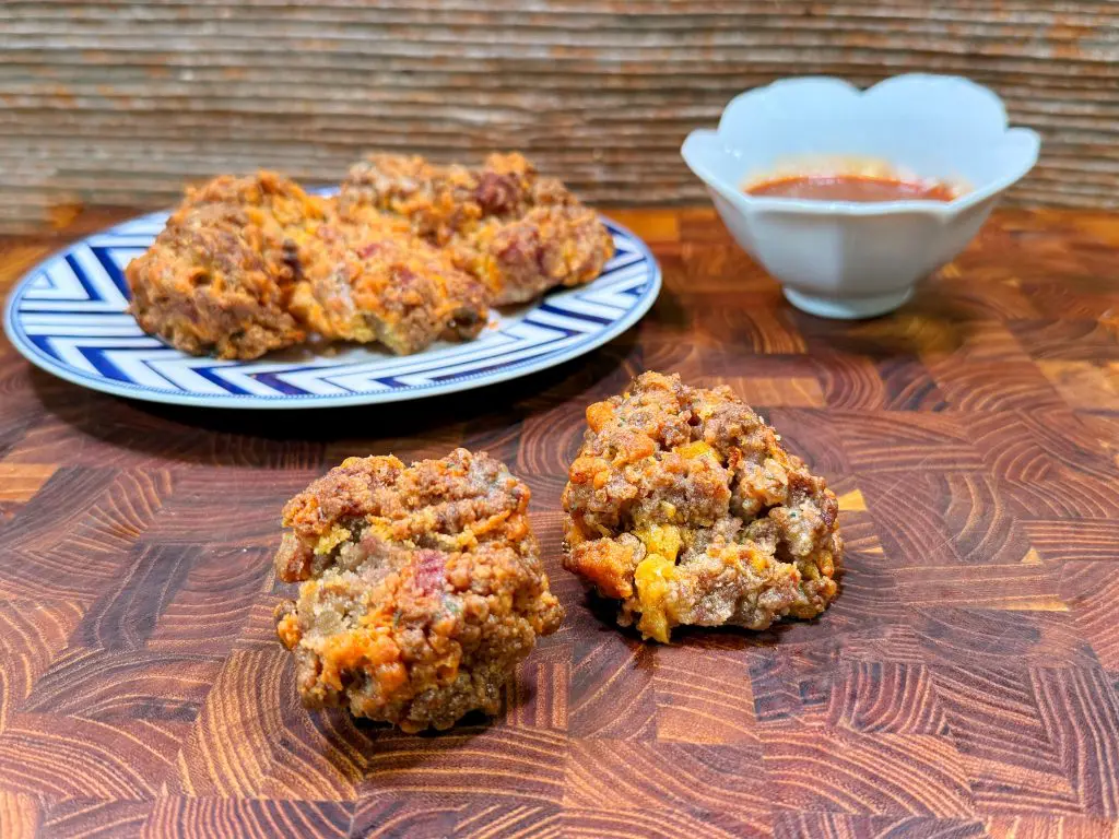 Two sausage balls sit on a wooden surface, with more sausage balls on a patterned plate and a small bowl of dipping sauce in the background.