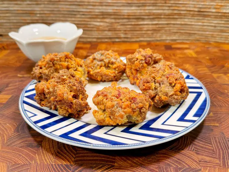 A blue and white plate with six golden-brown sausage balls sits on a patterned wooden surface, with a small white bowl containing dipping sauce in the background.