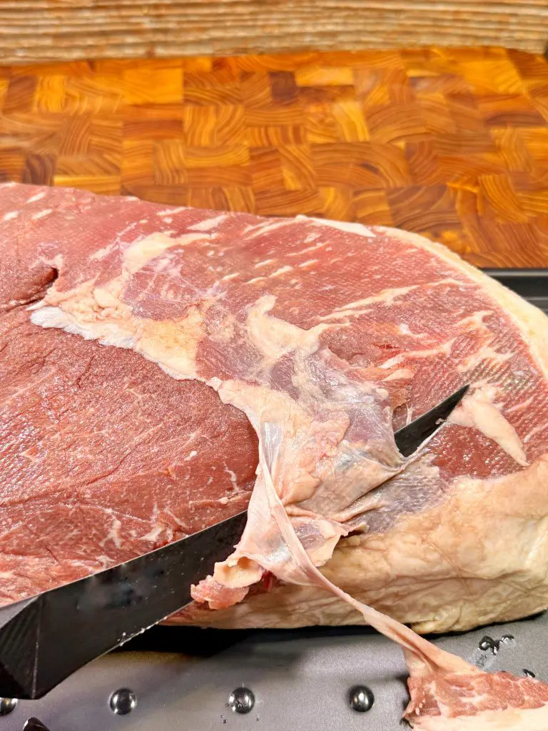 A close-up of a knife trimming silver skin from a raw cut of beef on a metal tray, with a wooden cutting board in the background.