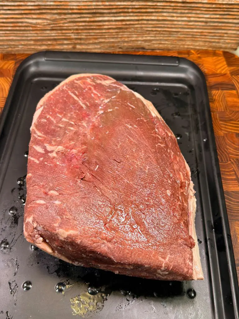 A large, raw beef brisket sits on a black roasting tray, ready for cooking. The meat is marbled with fat and has a reddish-brown color, with a textured wooden surface in the background. For a rump cap recipe
