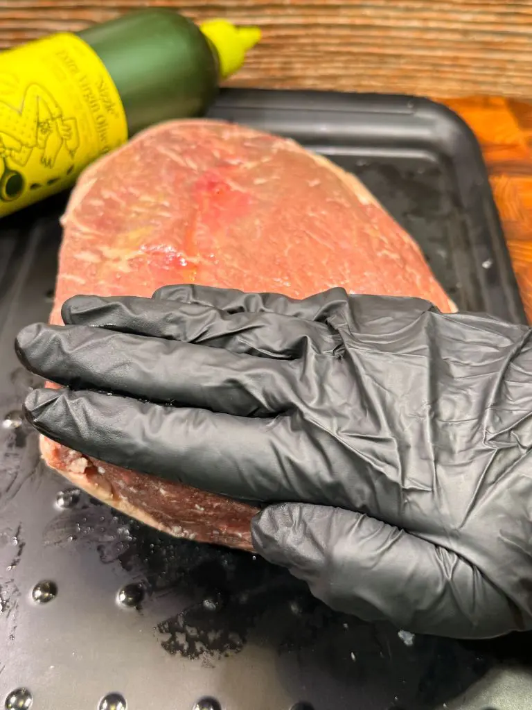 A hand wearing a black glove is pressing down on a raw steak placed on a black tray. A green bottle with a yellow nozzle is visible in the background.