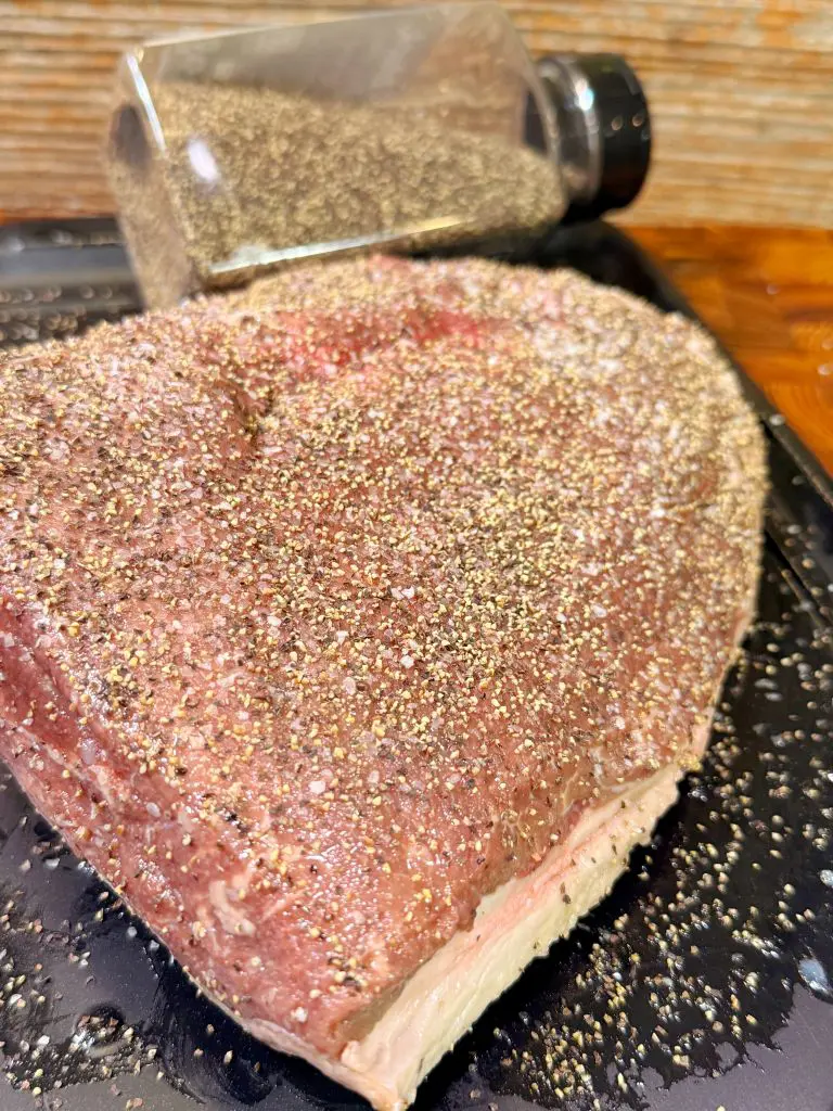 A large cut of raw beef, heavily coated with cracked black pepper, sits on a black tray. Behind it, a clear container of black pepper is tipped onto its side, spilling pepper onto the surface.