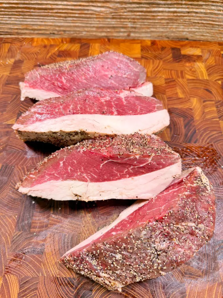 Four thick, raw beef steaks coated with coarse black pepper and seasoning are arranged on a wooden cutting board, with a rustic wood background visible at the top.