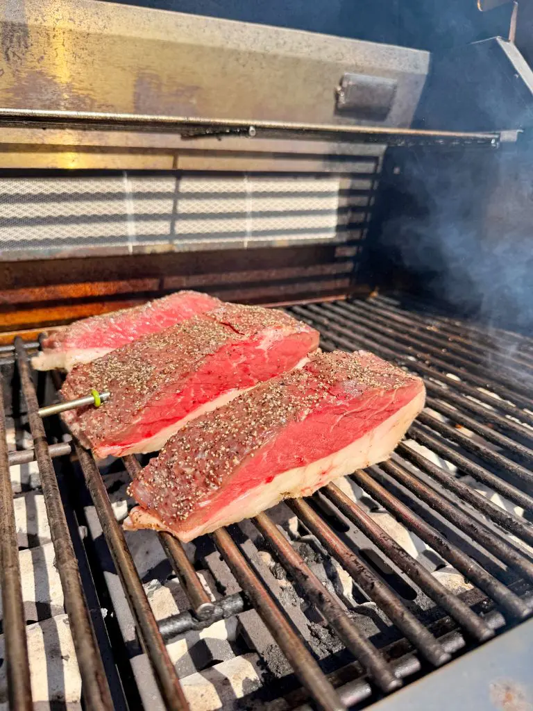Three thick, seasoned steaks are cooking on a grill with visible grill marks and smoke rising, suggesting they are being cooked outdoors on a barbecue.