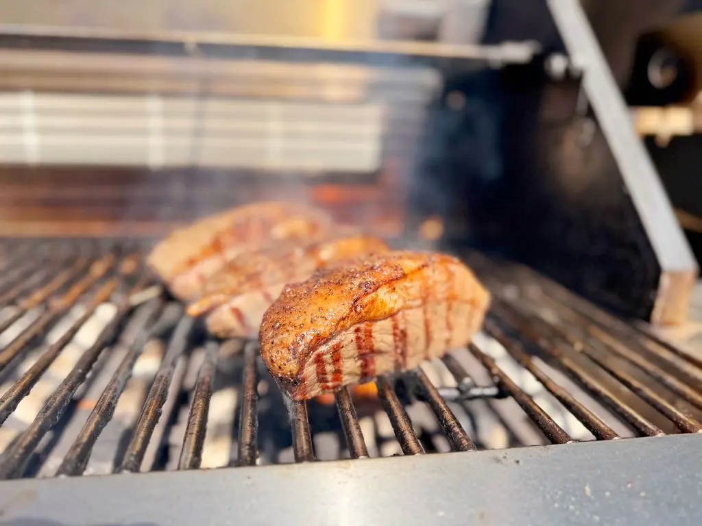 Three seasoned steaks grilling over an open flame on a barbecue, with visible grill marks and a slight smoky haze. The background is slightly blurred, emphasizing the sizzling meat in the foreground.