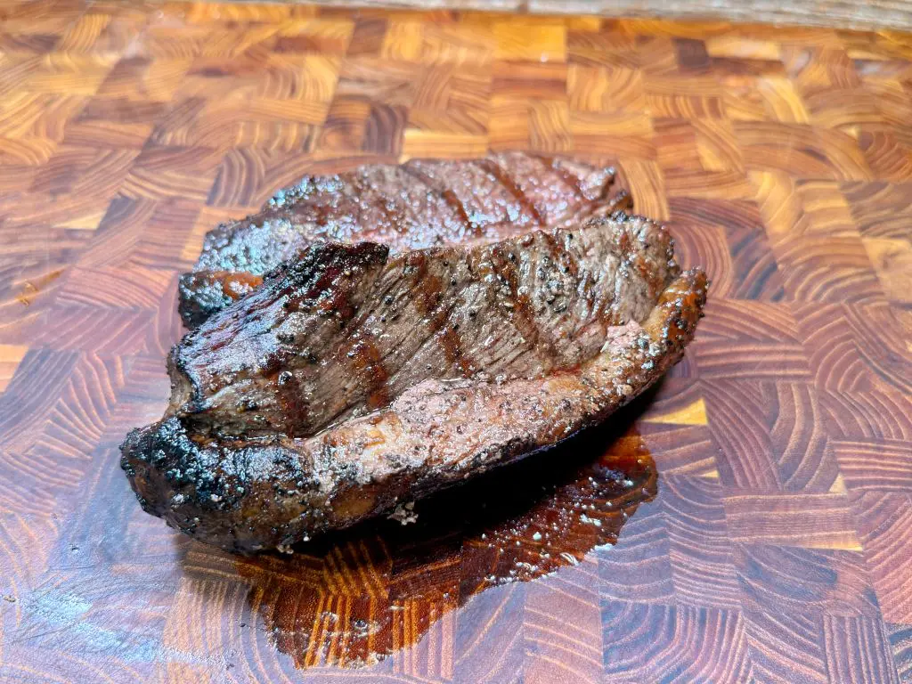 Two grilled steaks with visible grill marks rest on a wooden cutting board with a geometric pattern, showing a juicy, well-cooked exterior.