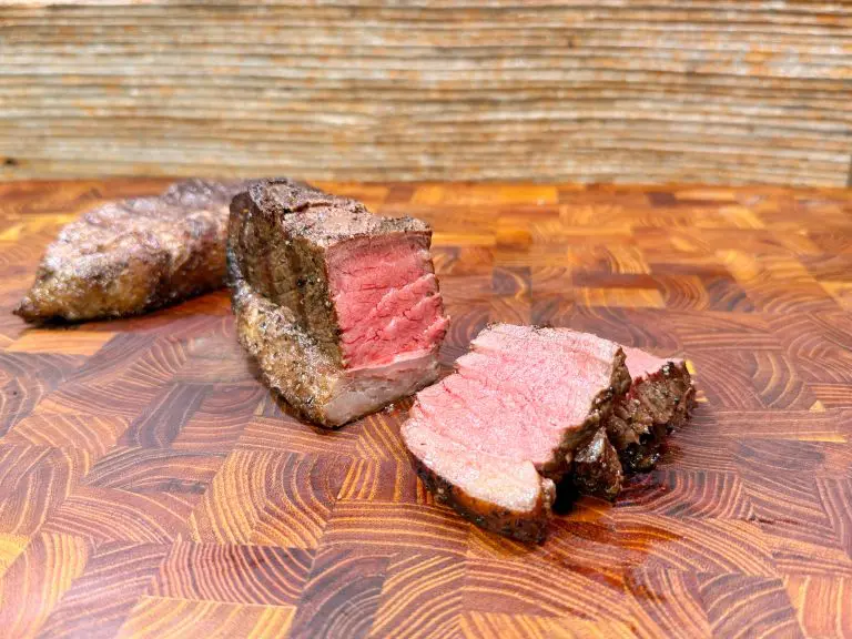 Two pieces of medium-rare steak, one whole and one sliced, rest on a patterned wooden cutting board, revealing a juicy pink interior and a seasoned brown crust.