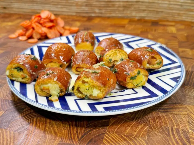 A plate with round, golden-brown stuffed bread rolls garnished with herbs. Sliced carrots are in the background on a wooden surface. The plate features a blue and white geometric pattern.
