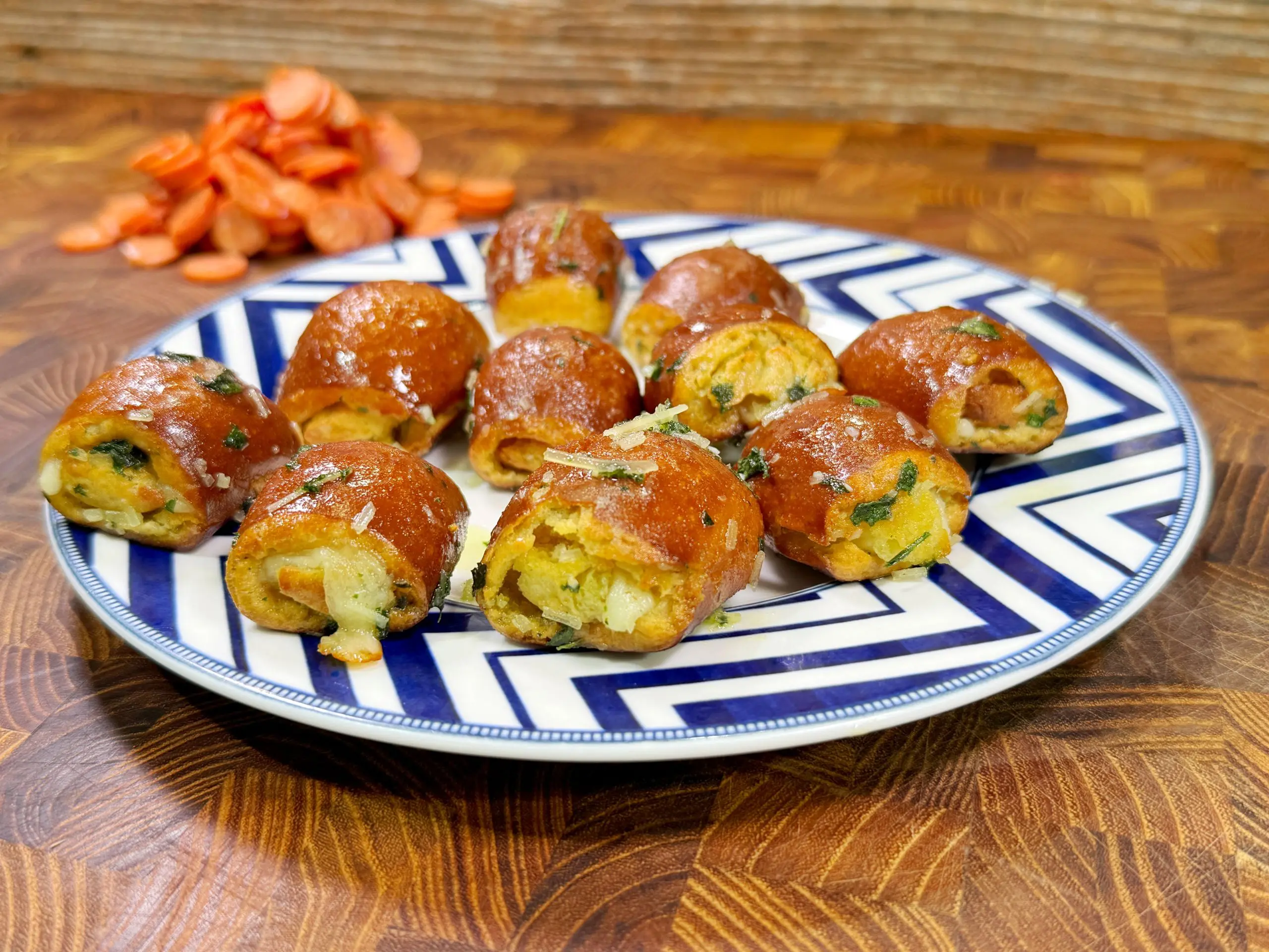 A plate with round, golden-brown stuffed bread rolls garnished with herbs. Sliced carrots are in the background on a wooden surface. The plate features a blue and white geometric pattern.