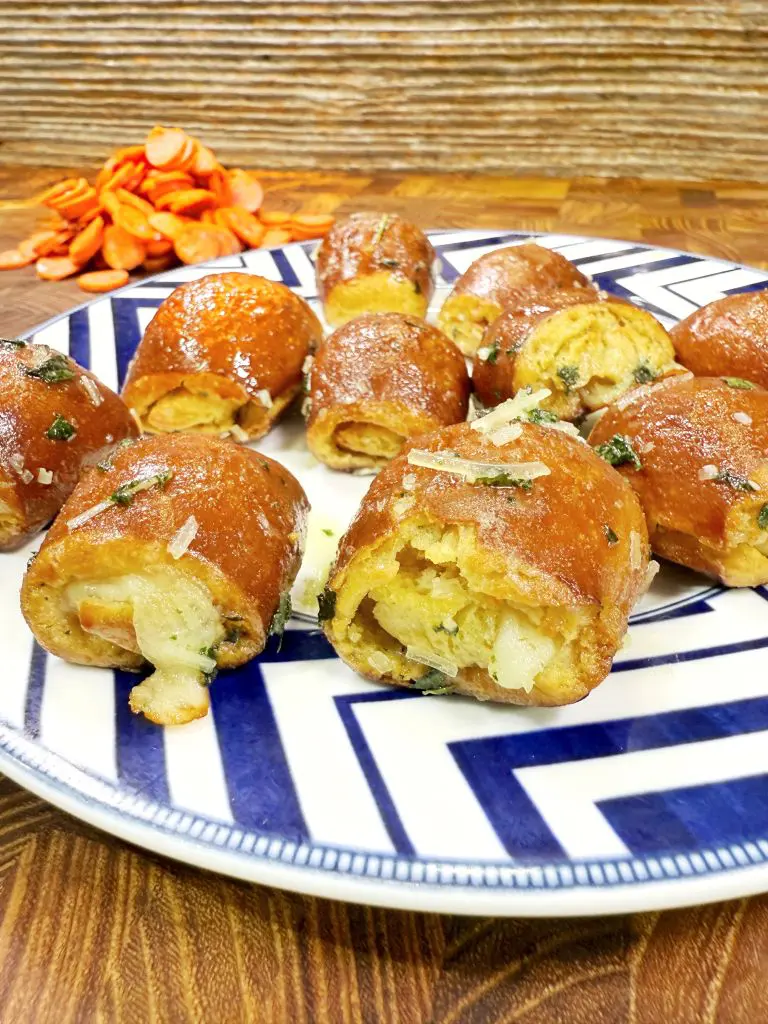 A plate of soft pretzel bites stuffed with cheese and herbs, topped with grated cheese and herbs. In the background, there is a small pile of sliced pepperoni on a wooden surface.