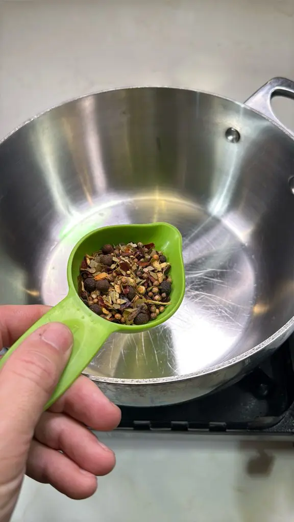 A hand holds a green measuring cup filled with mixed spices above an empty stainless steel pot on a stovetop, ready to pour the spices in.
