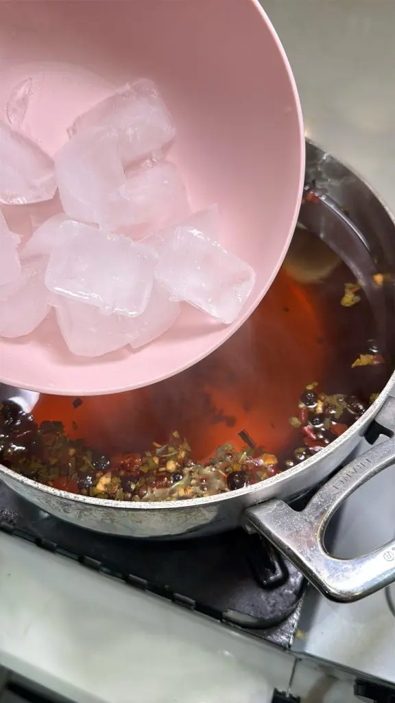 A person pours several ice cubes from a pink bowl into a pot of simmering broth with herbs and spices on a stovetop.