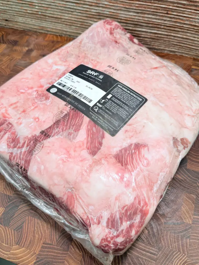 A large, vacuum-sealed cut of raw beef brisket with visible marbling sits on a wooden surface. The packaging has labels with product information and a barcode.