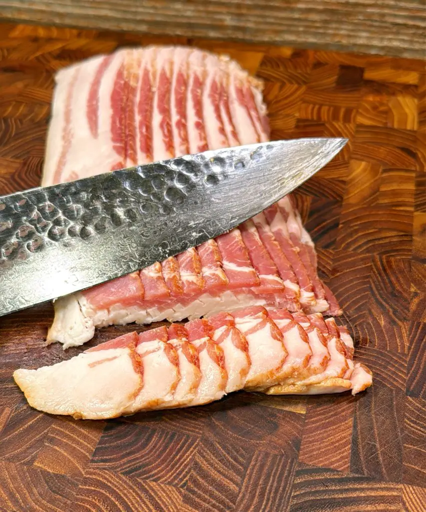 A sharp knife slicing thick strips of raw bacon on a wooden cutting board with a detailed, patterned surface.
