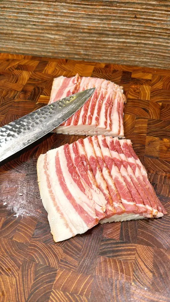 A knife rests on top of several slices of raw bacon arranged on a wooden cutting board with a textured pattern.