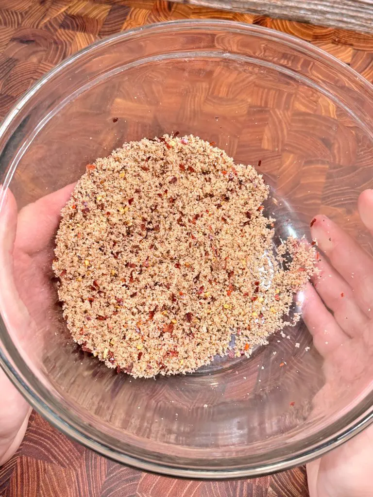 A person holds a glass bowl filled with a coarse, reddish-brown crumb mixture, possibly ground nuts or spices, over a wooden surface.