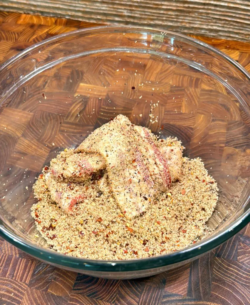 Raw fish fillets coated in a seasoned breadcrumb mixture sit in a clear glass bowl on a wooden countertop.