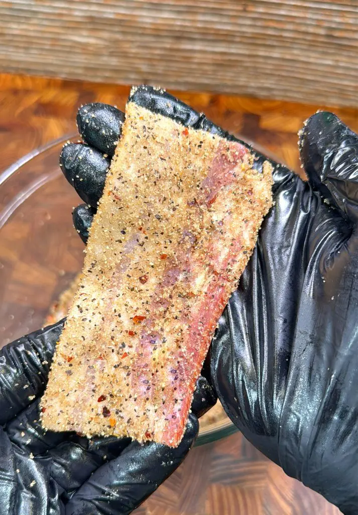 A gloved hand holds a strip of raw bacon coated with a mix of black pepper, crushed red pepper, and seasonings above a glass bowl. A wooden surface is visible in the background.