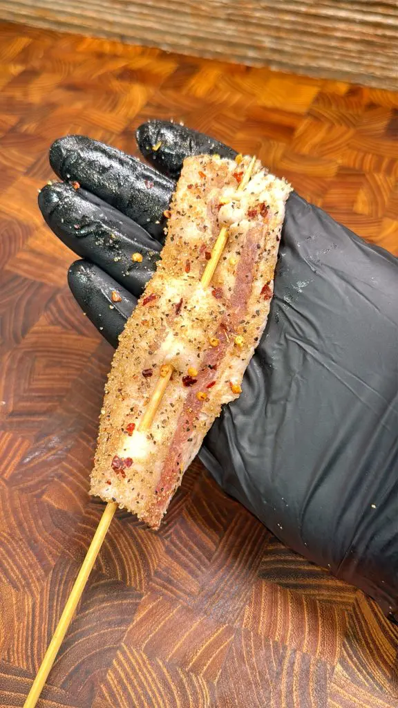 A gloved hand holds a skewer with a seasoned, grilled piece of meat, sprinkled with spices, over a wooden textured background. Recipe for billionaire bacon lollipops