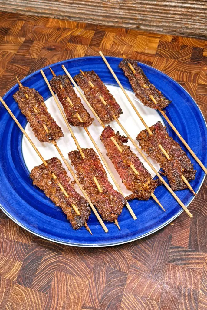 Several skewers of grilled beef suya, a West African spicy meat dish, are arranged on a blue and white plate, which is set on a wooden surface.