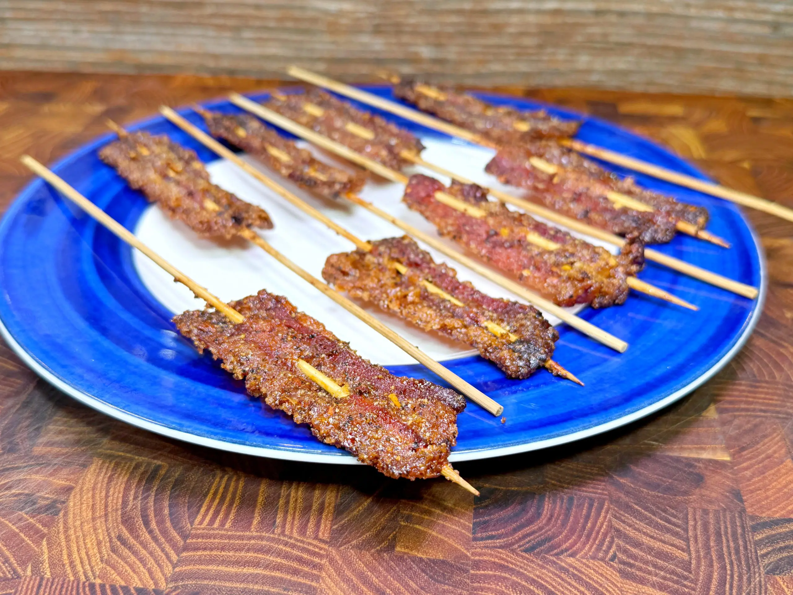 A blue and white plate holds several skewers of grilled, spiced meat arranged neatly on a wooden surface. The meat appears crispy and well-seasoned.