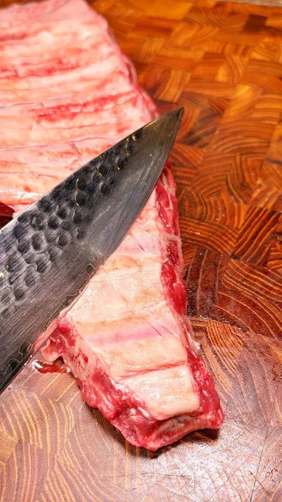 A close-up of a large knife scoring raw beef ribs on a patterned wooden cutting board. The meat and knife are in sharp focus, highlighting the marbling and texture.