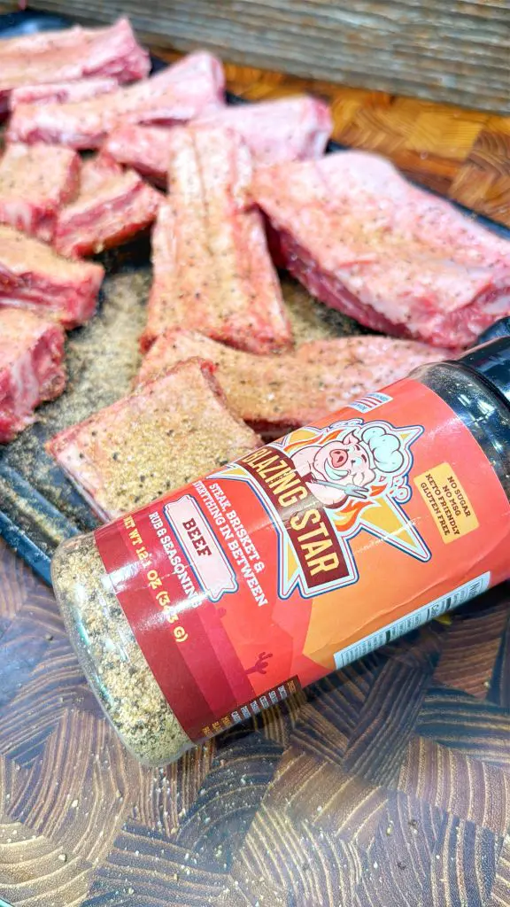 Several raw beef short ribs are seasoned and arranged on a cutting board. In the foreground, a bottle of All-Star BBQ seasoning labeled Beef rests on its side, partially covering some of the meat.