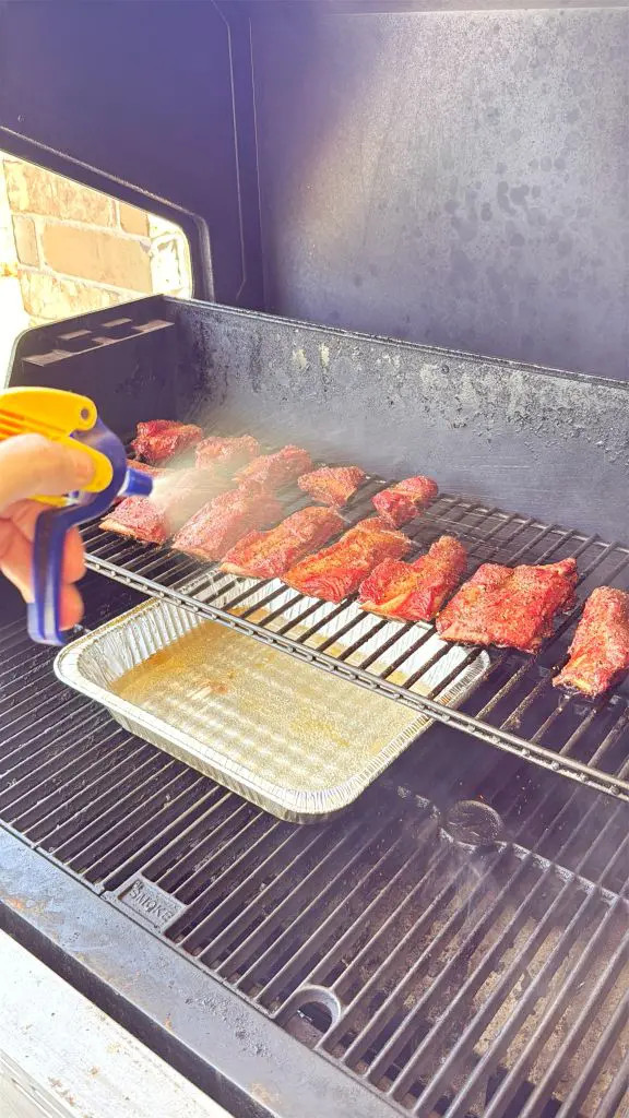 A person sprays liquid onto rows of seasoned meat cooking on a grill, with a disposable tray placed underneath to catch drippings. The scene is outdoors, next to a brick wall.