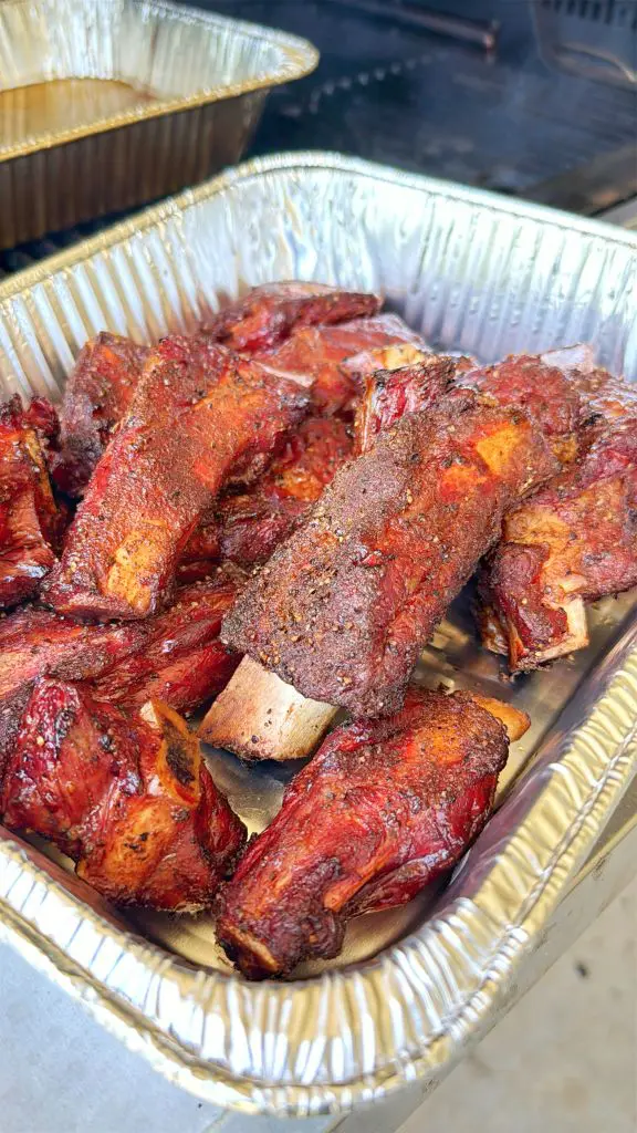 A foil tray filled with several pieces of smoked beef ribs with a dark, seasoned crust, resting on a metal surface, likely at a barbecue or outdoor cookout.