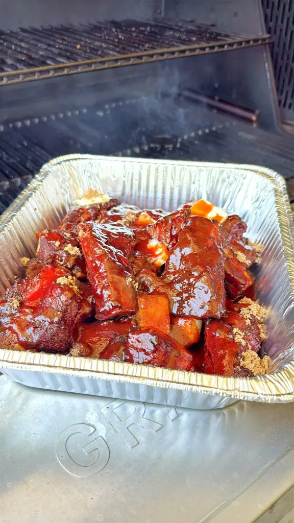 A foil tray filled with glazed, saucy beef ribs sits on the surface of a barbecue grill, with smoke rising from the grill in the background.