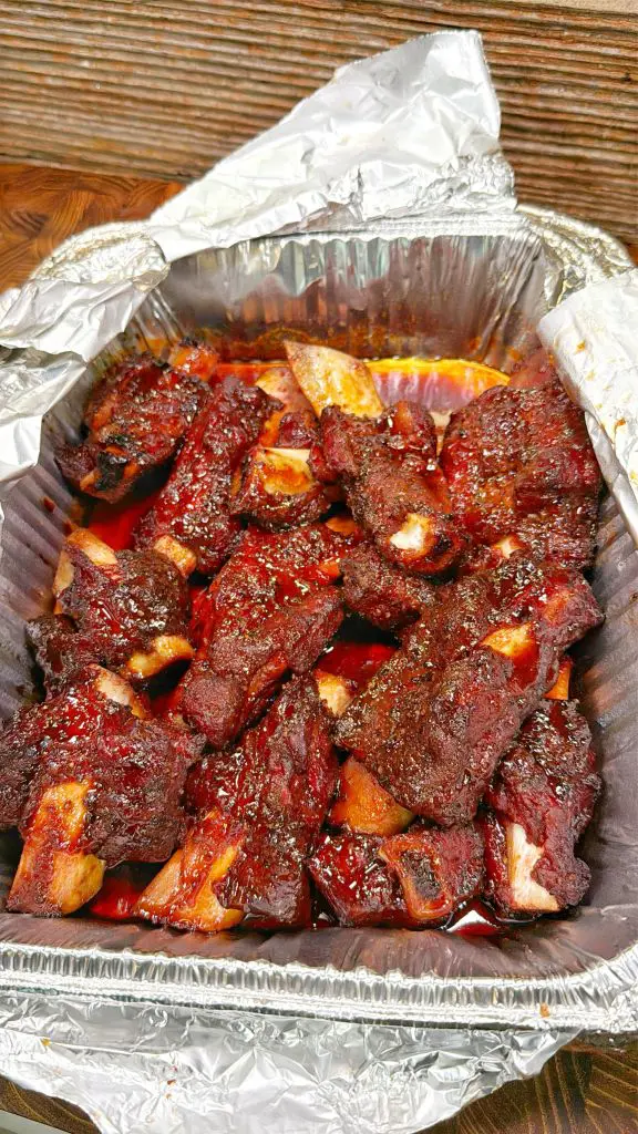 A foil tray filled with barbecue beef ribs covered in a rich, dark sauce, with the foil partially unwrapped and juices pooled at the bottom.