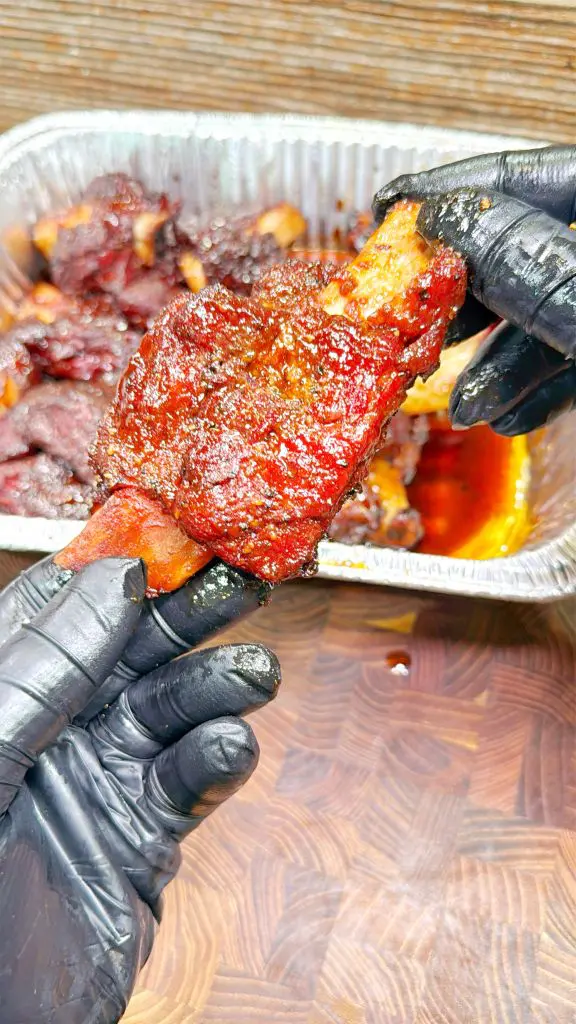 A person wearing black gloves holds a saucy, cooked meat rib over a wooden surface, with a foil tray containing more ribs and sauce in the background.