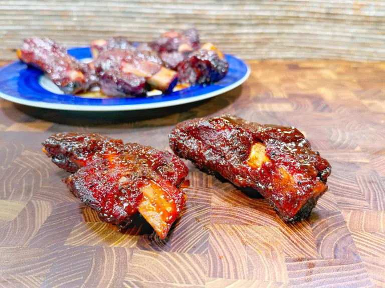 Two glazed, barbecued beef ribs on a wooden cutting board, with a blue plate of more ribs in the background. The ribs are coated in a shiny, dark red sauce.