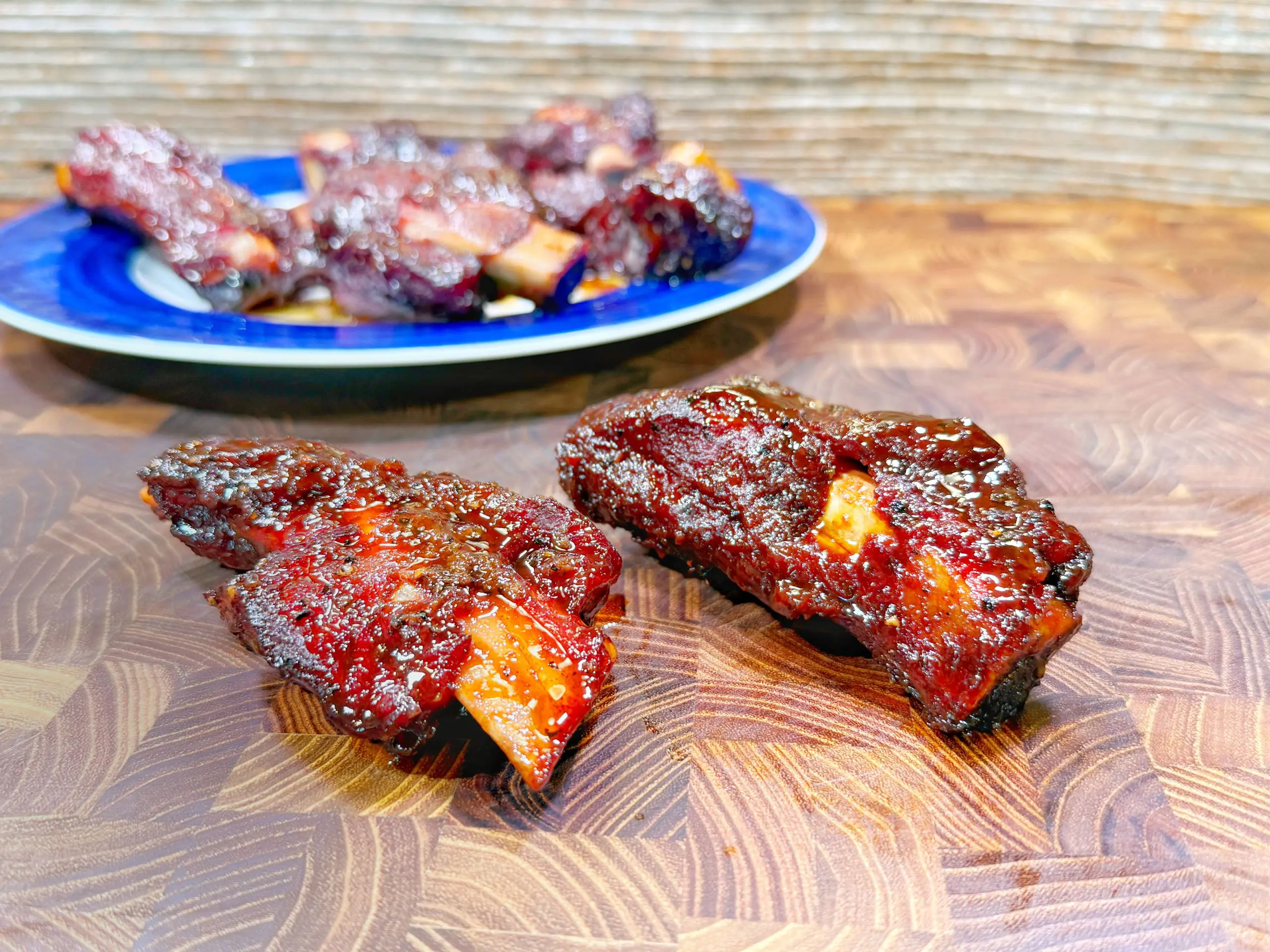 Two glazed, barbecued beef ribs on a wooden cutting board, with a blue plate of more ribs in the background. The ribs are coated in a shiny, dark red sauce.